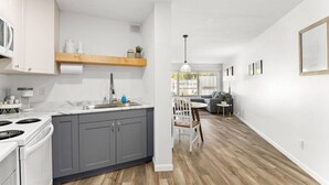 Cocina moderna con gabinetes grises, estufa blanca y encimera de madera. Zona de comedor con mesa y sillas, y sala de estar con sofá gris y lámpara de pie.