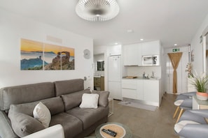 A modern living room with a sofa, a coffee table, and a dining area with chairs. There is a kitchen in the background with white cabinets and appliances.