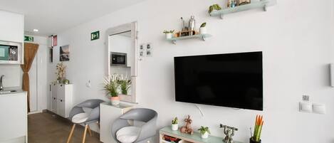 A modern hotel room with a flat-screen TV, a dining area with chairs, a kitchen area, and decorative shelves.