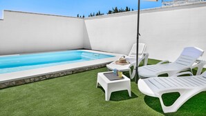 Uma área de piscina com uma piscina branca, duas espreguiçadeiras brancas, uma mesa pequena com um chapéu e uma xícara, e um grande guarda-sol.