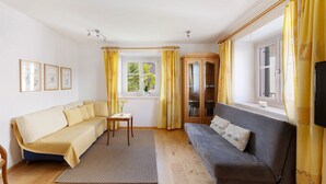 A living room with a sofa, a small table, a bookshelf, and a window with curtains. - Apartment 'Kreutnerbinderhof' with Mountain View, Private Terrace and Wi-Fi (Terlano)