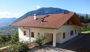 A two-story house with a red-tiled roof, white walls, and a view of mountains.