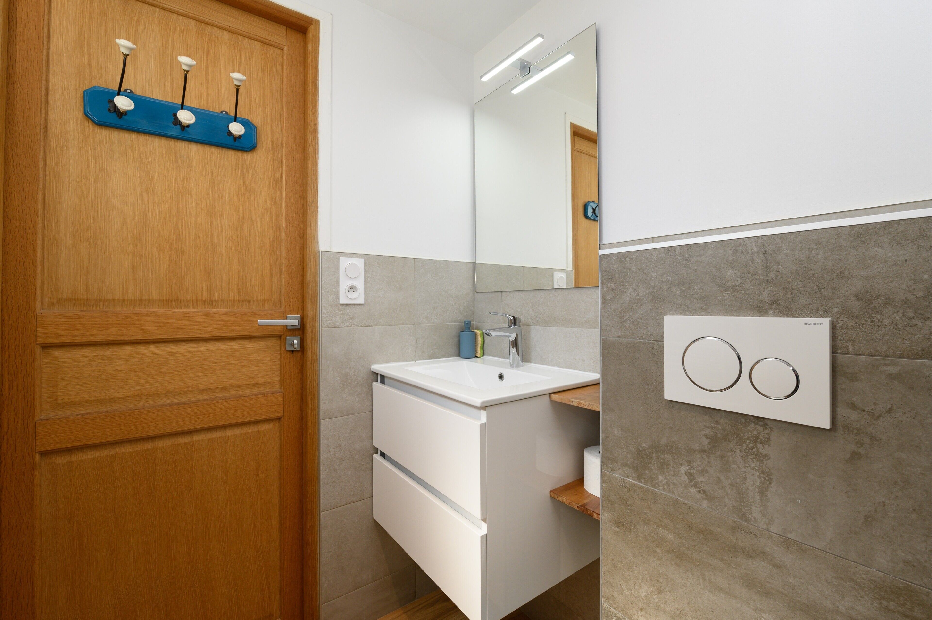 Un baño con puerta de madera, un lavabo blanco con espejo y una lámpara de pared.
