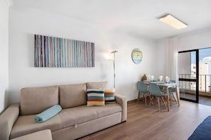 A modern living room with a sofa, a dining table with chairs, a floor lamp, and a large window with a view. - Apartment 'Seaside Heaven - Praia Da Rocha' with Mountain View, Shared Pool and Wi-Fi (Portimão)