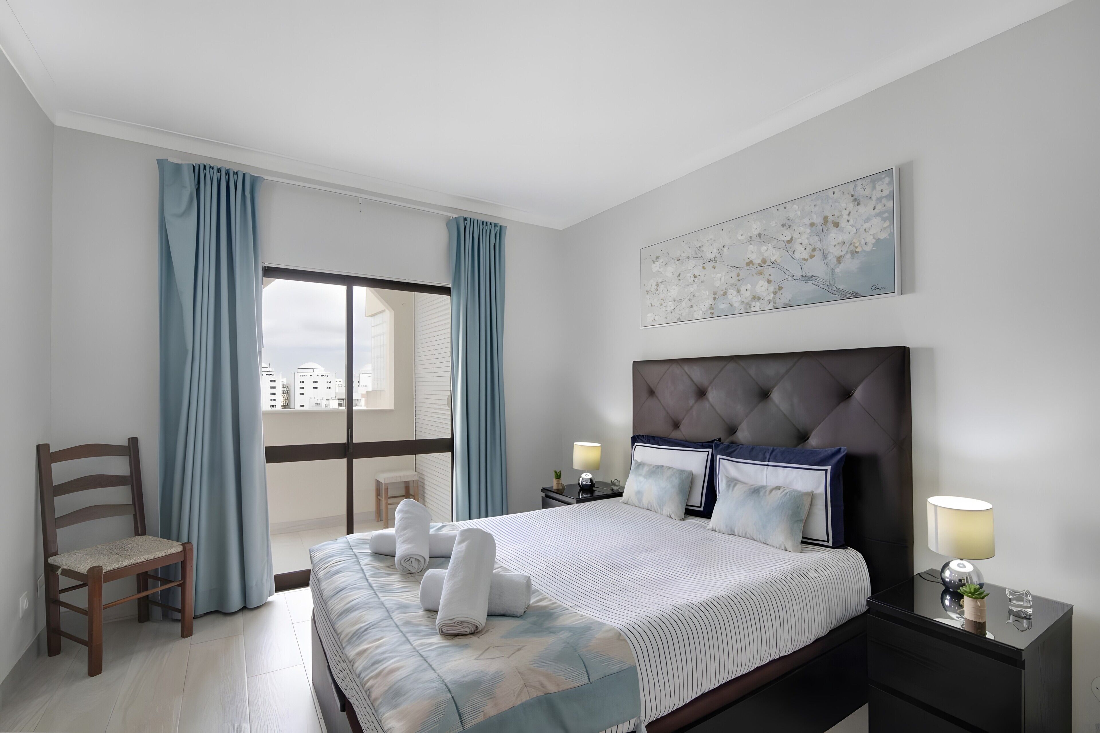 Una habitación de hotel con cama, una silla, una mesita de noche, una ventana con cortinas y una obra de arte colgada en la pared.