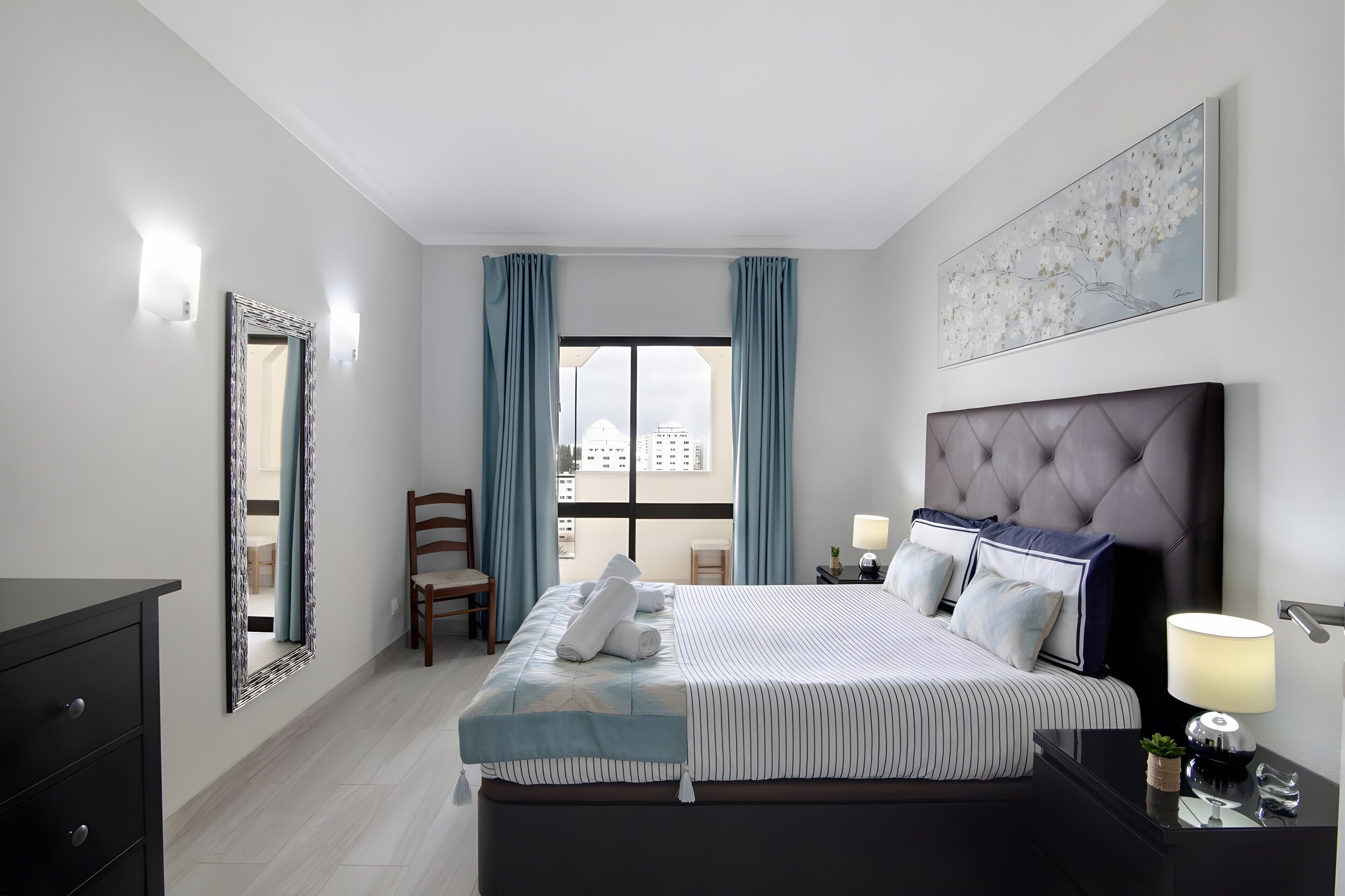 Una habitación de hotel con cama, una silla, escritorio, lámpara y ventana con cortinas.