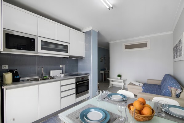 A modern kitchen with built-in appliances, a dining area with a table set for two, and a living room with a sofa and a small plant.