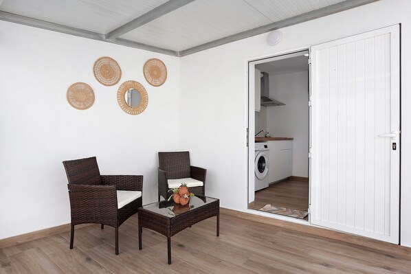 Une pièce comprenant deux fauteuils en osier, une table avec un plateau en verre sur laquelle est posée une plante, et une porte blanche donnant sur une buanderie.