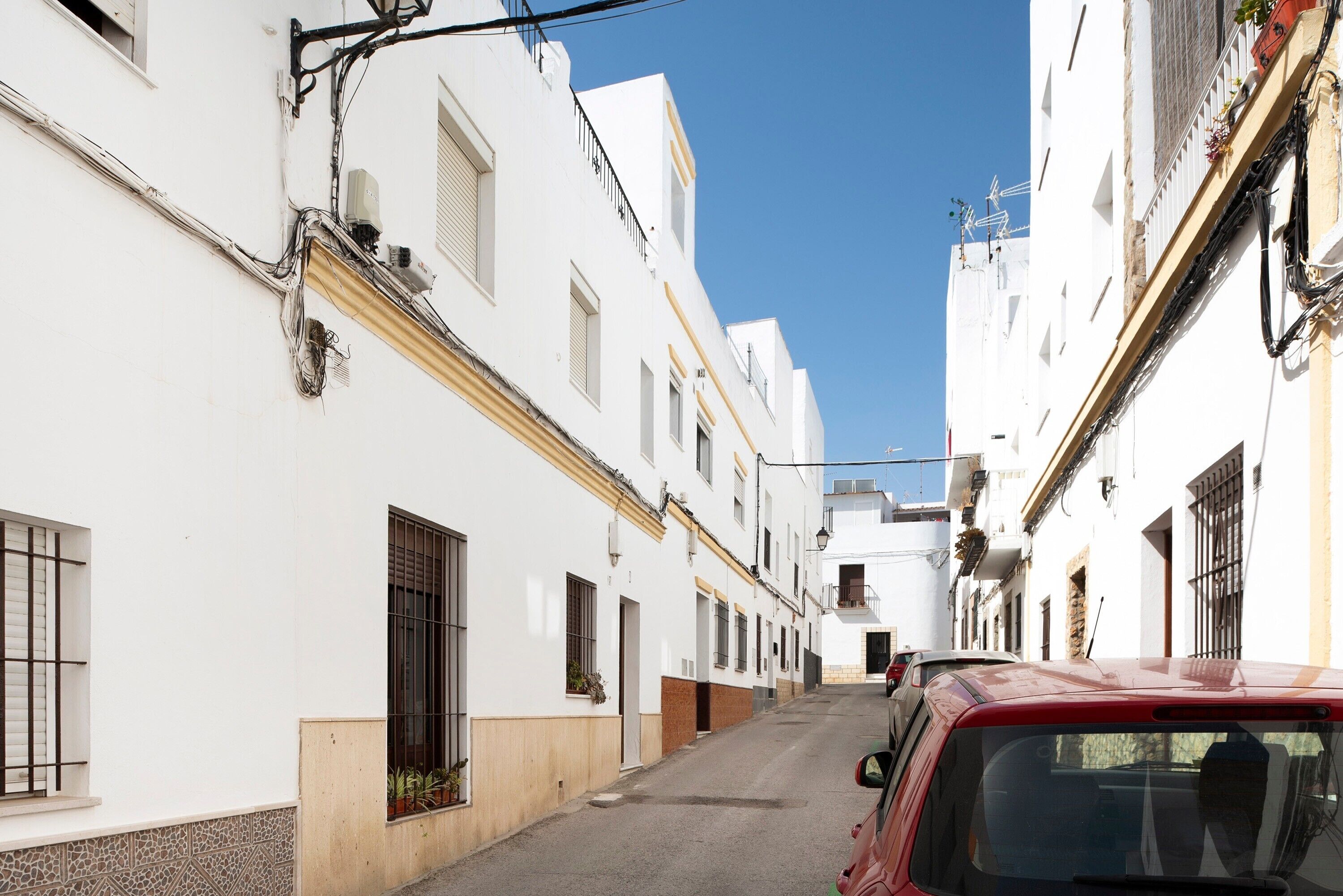 A narrow street with white buildings, a red car parked on the right, and a clear blue sky.