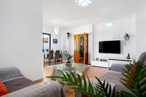 A modern living room with a glass coffee table, a flat-screen TV, and a dining area in the background.