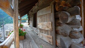 Un chalet rustique en rondins de bois, doté d’un porche en bois, d’un toit en pierre et offrant une vue sur un paysage montagneux.