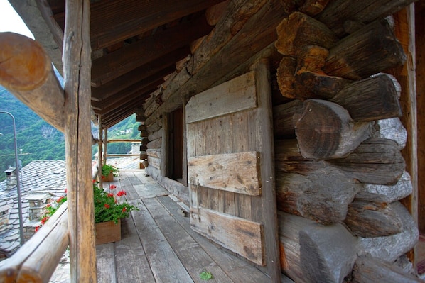 A rustic log cabin with a wooden porch, stone roof, and a view of a mountainous landscape.