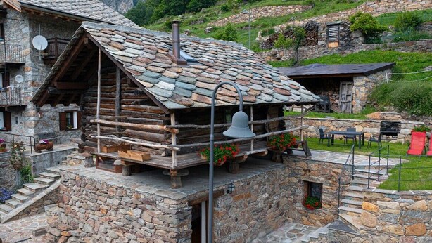 Une cabane rustique en rondins de bois, avec des fondations en pierre, un balcon agrémenté de plantes en pot et une cloche.