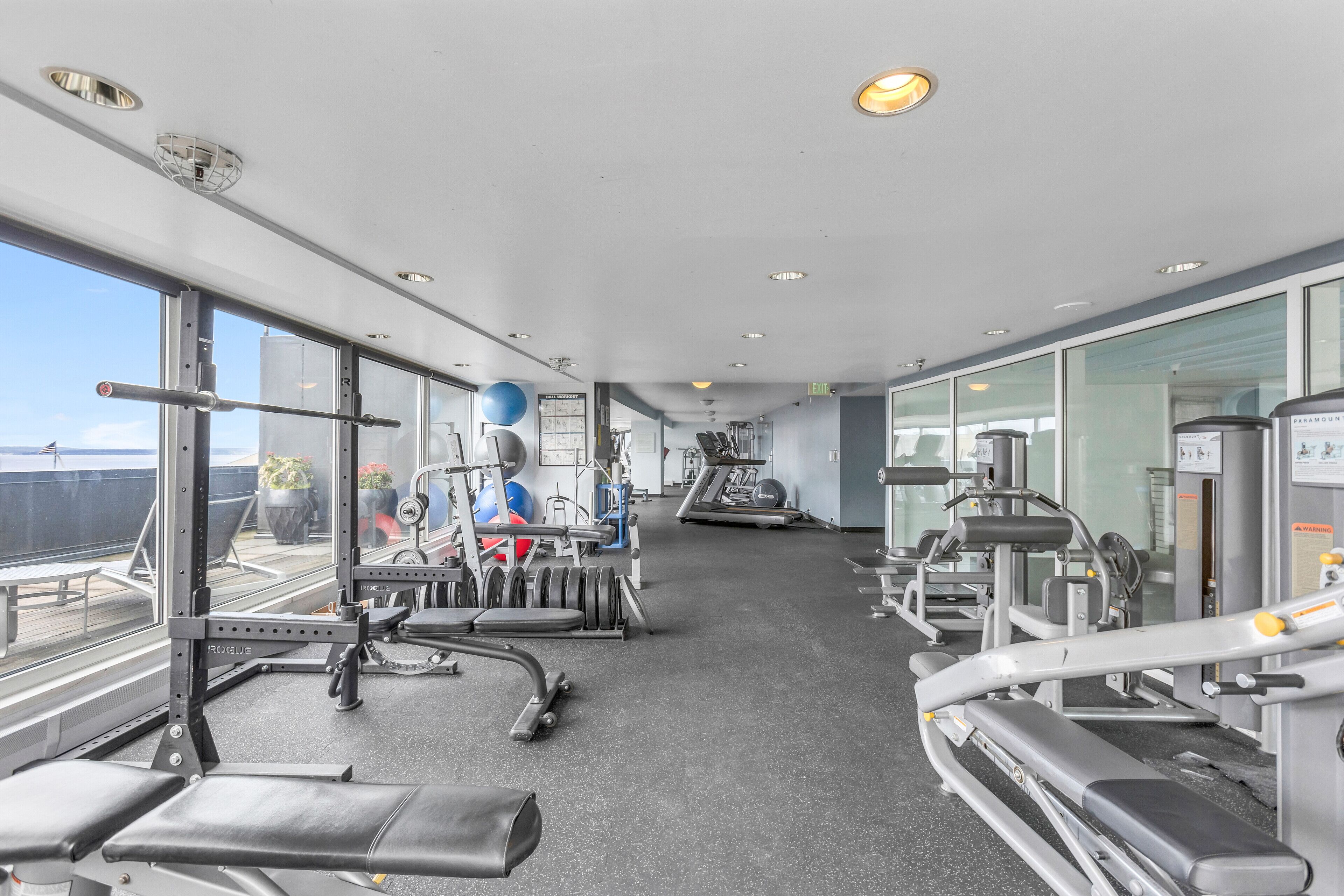 Ett gym med olika typer av träningsutrustning, stora fönster och utsikt över staden.