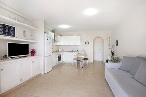 Une cuisine moderne avec des meubles blancs, une table à manger avec des chaises, un canapé gris et un téléviseur posé sur le comptoir.