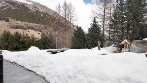 Un espace extérieur enneigé, avec un banc en pierre, une structure en bois et une chaîne de montagnes en arrière-plan.