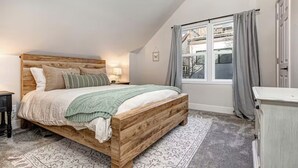 Quarto com cama de madeira, mesa de cabeceira, luminária, janela com cortinas e radiador.