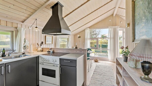 Une cuisine équipée d’une cuisinière, d’un four et d’une hotte noire. On y trouve une fenêtre avec des rideaux, un évier et un coin repas avec des chaises et une table.