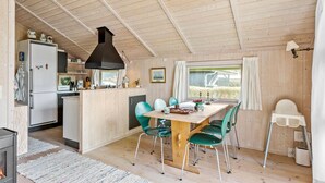 L’intérieur chaleureux d’un chalet avec une table à manger en bois, des chaises de couleur turquoise, un coin cuisine équipé d’un réfrigérateur et d’une cuisinière, et une grande fenêtre offrant une vue sur l’extérieur.