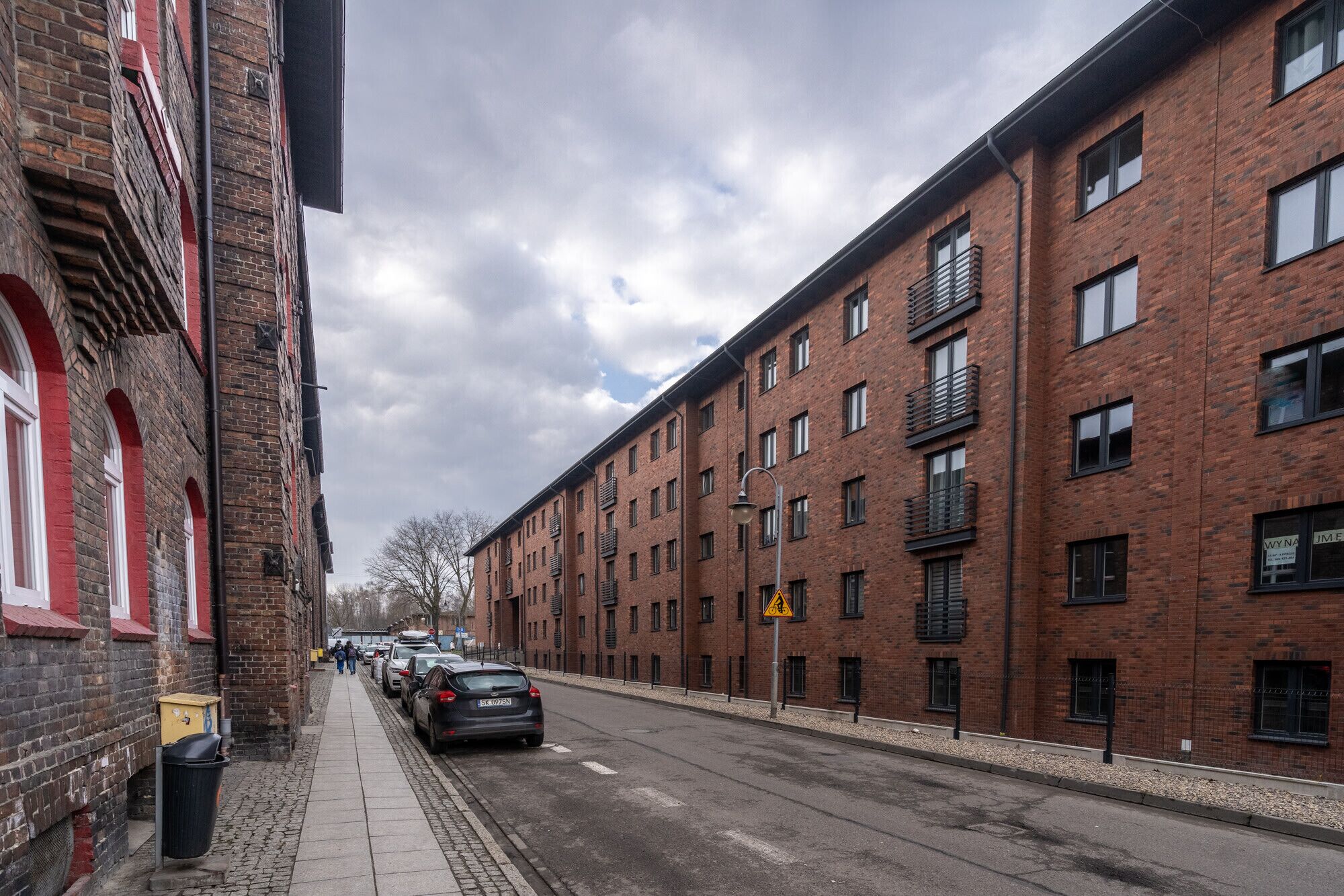 A street view with brick buildings, a pedestrian sidewalk, and parked cars.