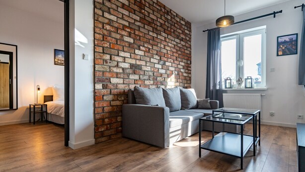A modern living room with a brick accent wall, a grey sofa, a glass coffee table, and a view of the city through a window.