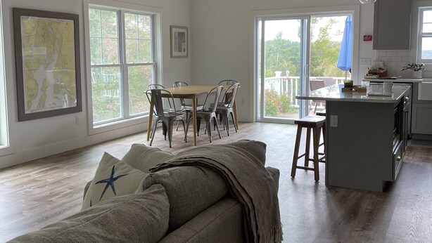 A modern living room with a sofa, dining table, and chairs. There is a kitchen area with a counter and cabinets. Large windows provide natural light and views of the outdoors.