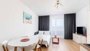 A modern hotel room with a bed, a dining table with chairs, a small red side table, a television, and a painting on the wall.