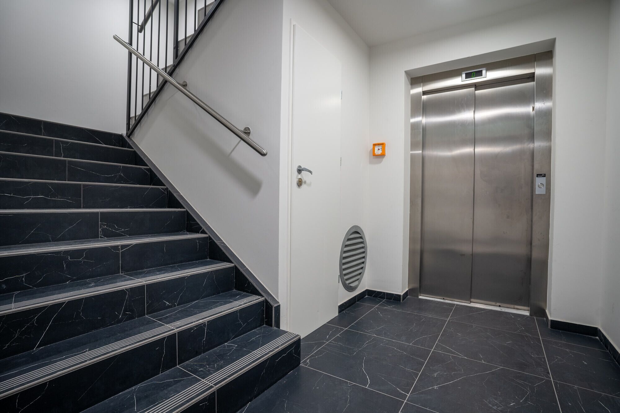 Una escalera con escalones de mármol negro y barandilla de metal, que conduce a una puerta blanca con cerradura y detector de incendios.