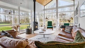 A spacious living room with a large fireplace, a green armchair, a leather sofa, and a view of the outdoors through large windows. - 4 bedroom pet friendly home in Strandby (Strandby)
