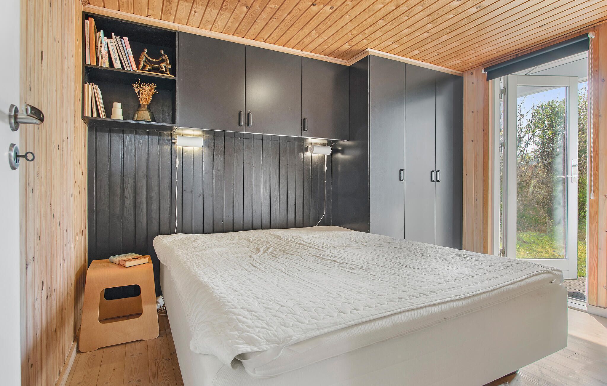 Une chambre à coucher comprenant un lit, une armoire en bois, une étagère avec des livres et des objets décoratifs, et une porte-fenêtre coulissante donnant sur l’extérieur.
