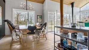 A spacious room with a wooden ceiling, large windows, a dining table with chairs, and a kitchen area with shelves. - 4 bedroom pet friendly home in Strandby (Strandby)