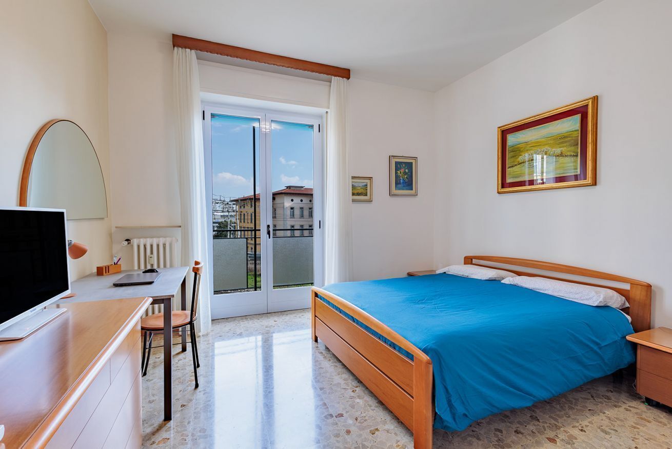 Un dormitorio con cama, escritorio, silla, televisor y ventana con vista a edificios.