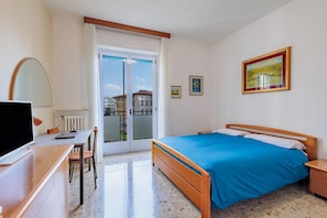 A bedroom with a bed, a desk, a chair, a TV, and a window with a view of buildings.