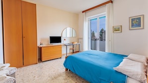 Un dormitorio con cama, escritorio, televisor, armario y balcón.