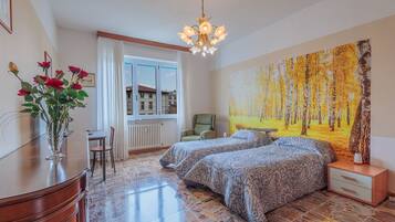 Un dormitorio con dos camas, un escritorio, una silla, un radiador, una ventana con cortinas y un cuadro en la pared.