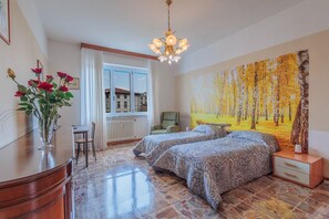 Un dormitorio con dos camas, un escritorio, una silla, un radiador, una ventana con cortinas y un cuadro en la pared.