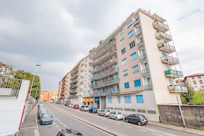 A multi-story residential building with balconies, parked cars along the street, and a clear sky.