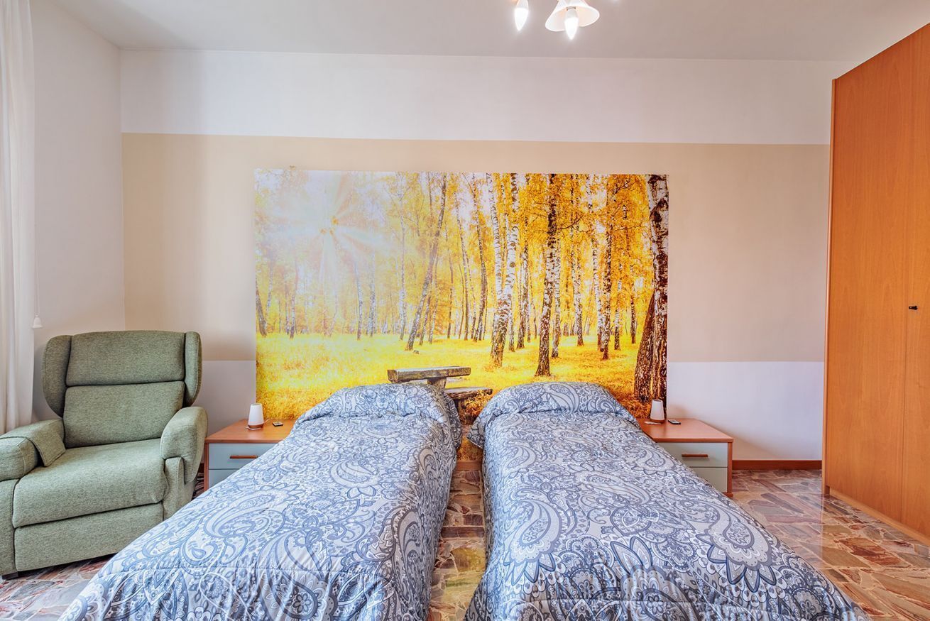Ein Zimmer mit zwei Betten, einem grünen Sessel, einem Waldgemälde und einer Holztür.