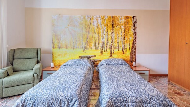 Una habitación con dos camas, un sillón verde, un cuadro de un bosque y una puerta de madera.