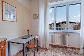Una habitación con escritorio, silla, radiador y ventana con vistas a edificios.