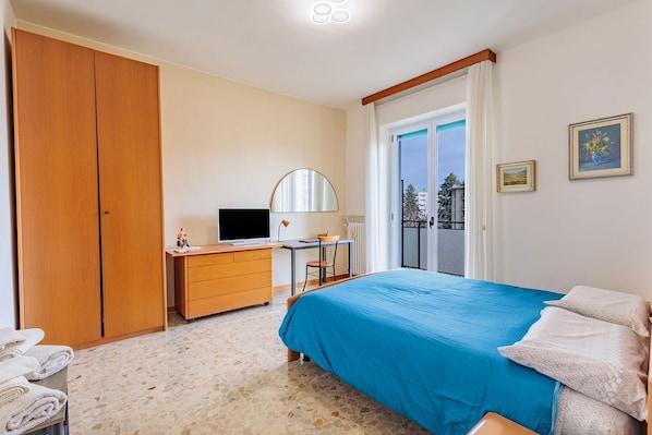 A bedroom with a bed, a desk, a television, a wardrobe, and a balcony.