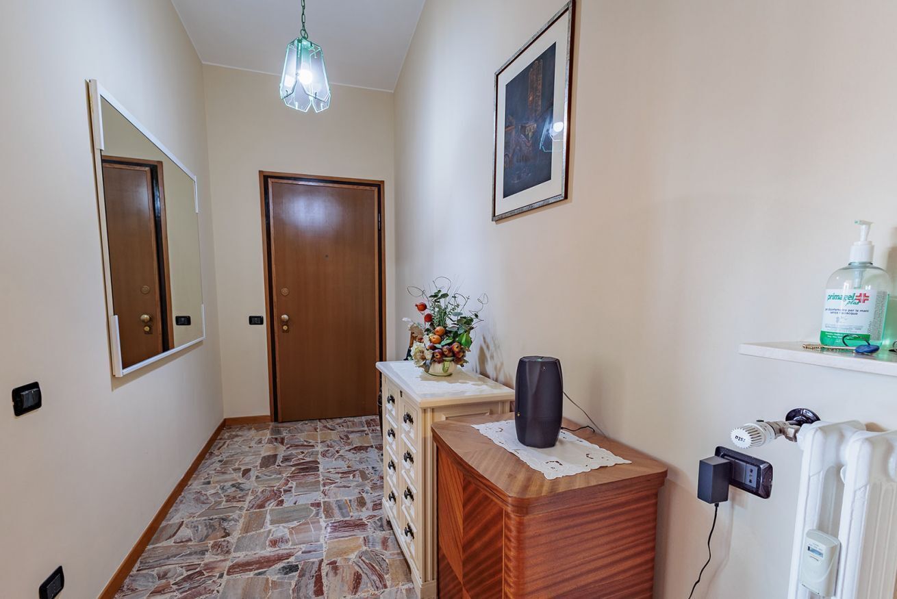 Un pasillo con un aparador de madera, un espejo, un cuadro enmarcado, un jarrón con flores y un radiador.