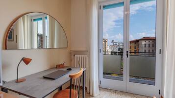Habitación con escritorio, silla y un espejo grande. Hay una puerta corrediza de vidrio que da a un espacio exterior con vistas a edificios.