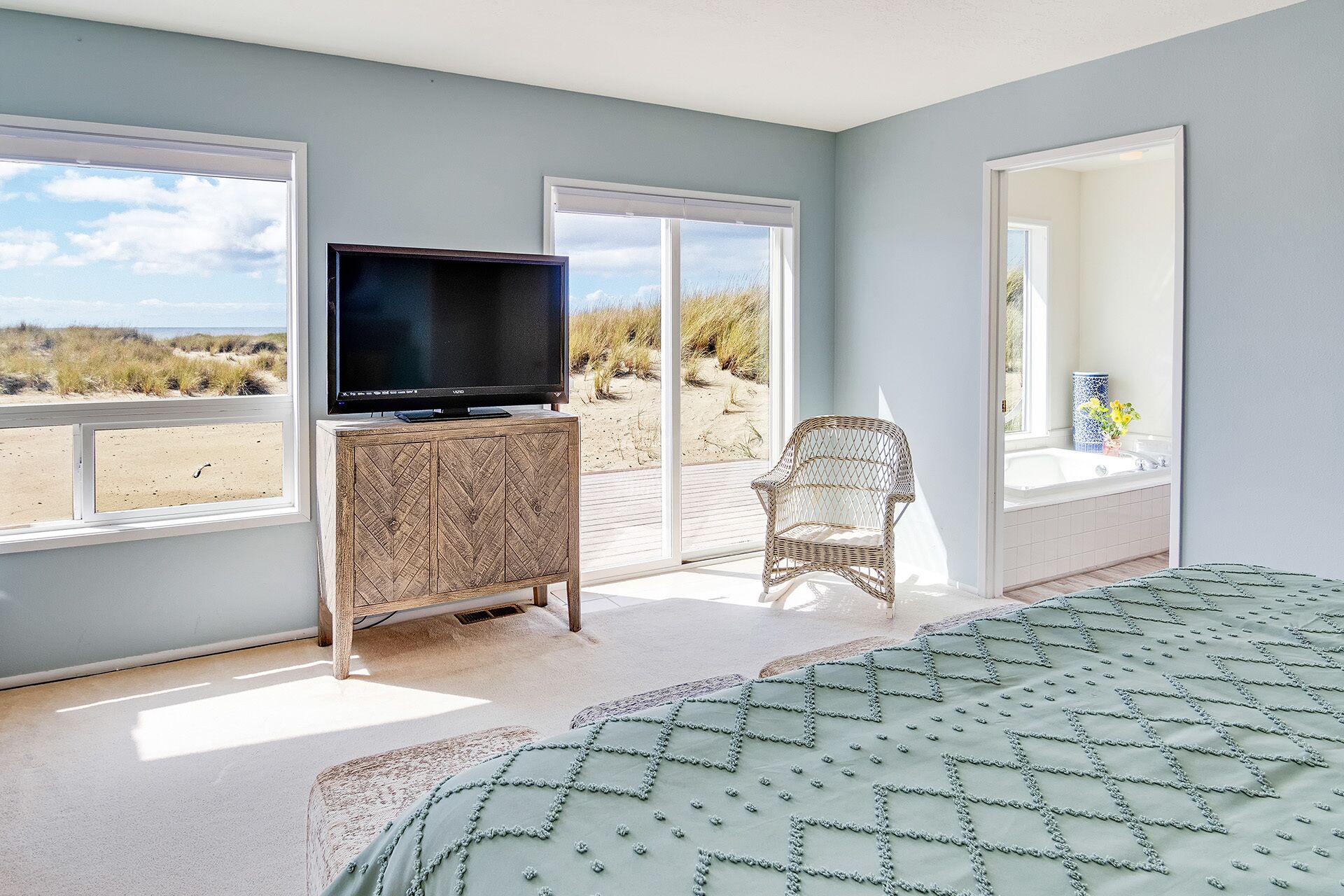 A bedroom with a large window, a television on a wooden cabinet, a wicker chair, and a view of the beach.