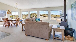A spacious living room with a large beige sofa, wooden dining table, and chairs. There are large windows offering views of the beach. A fireplace is visible on the right side. - Ocean Front with Views! Bridge Views! Sandy Beach! (Waldport)