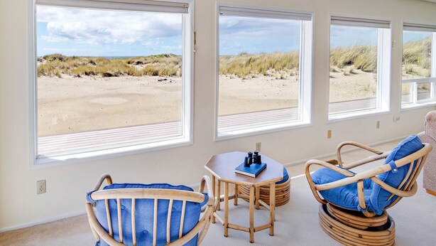 A room with large windows overlooking a beach, featuring wicker chairs and a small table with binoculars. - Ocean Front with Views! Bridge Views! Sandy Beach! (Waldport)