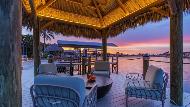 A covered outdoor seating area with chairs and a table, overlooking a body of water at sunset. - GULF ACCESS & BIG WATER VIEW!, Heated Saltwater Pool  and Spa -  Villa Manatees Crossing - Roelens (Cape Coral)