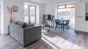 Una sala moderna con un sofá gris, un comedor con sillas azules y una cocina con armarios blancos.