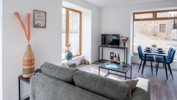 Una sala moderna con un sofá gris, una mesa de centro, un televisor sobre un mueble bajo, y un comedor con sillas azules y una mesa.
