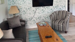A living room with a black sofa, a patterned armchair, a wooden coffee table, and a wall-mounted TV. - Tiny house on an orchard (Oliver)